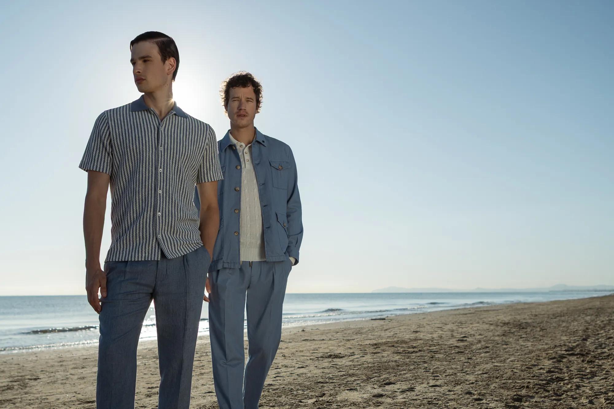 Two men in summer clothing walking along a beach coastline under a clear sky