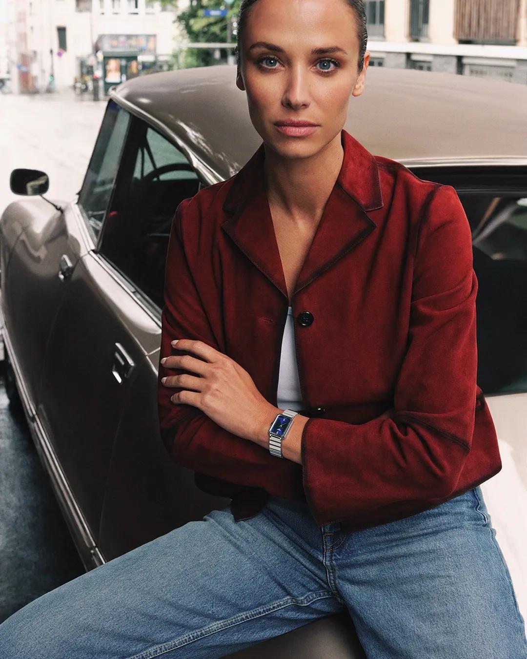 Woman in a red jacket and jeans sitting in front of a classic car wearing a wristwatch