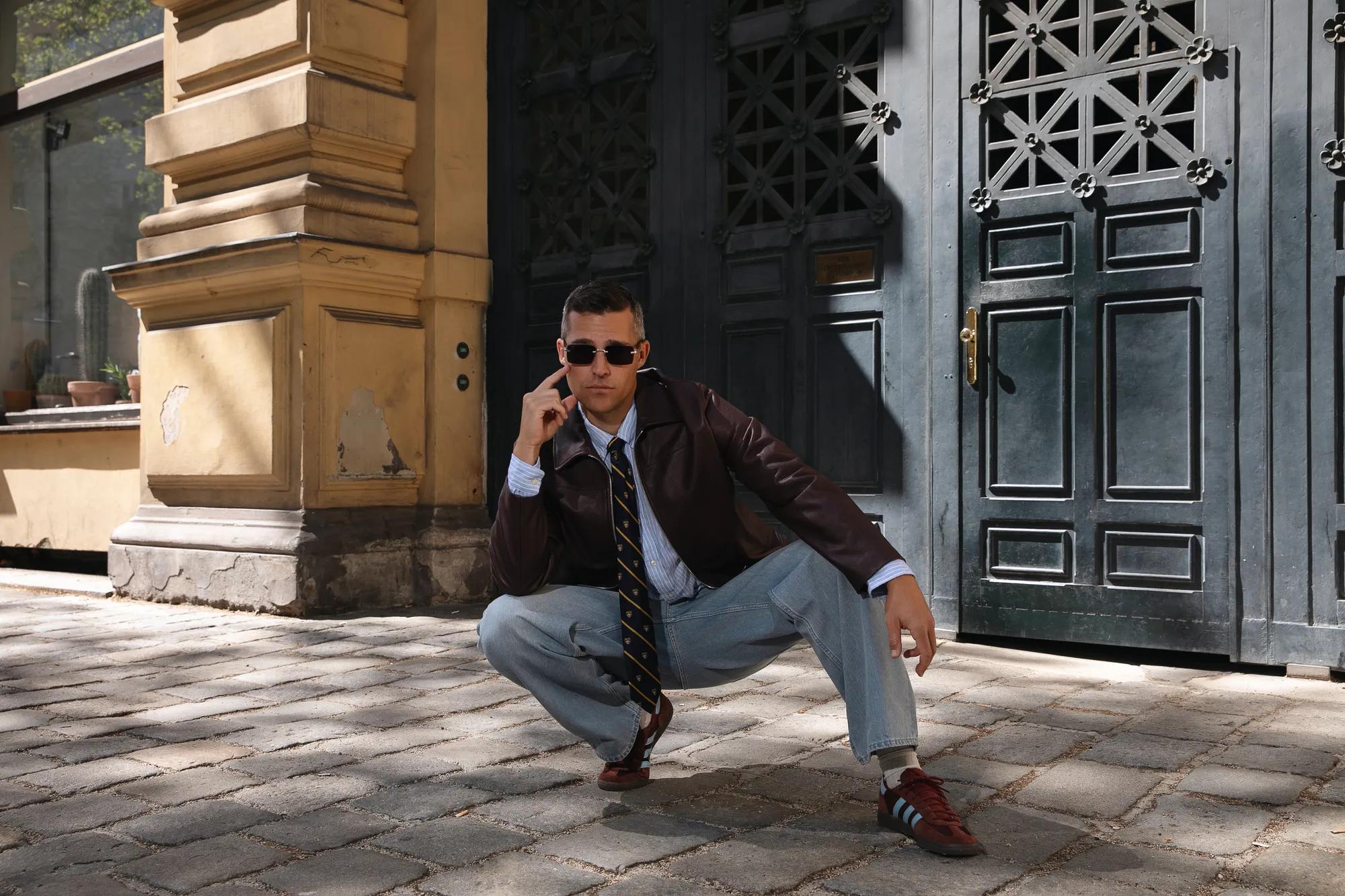 Stylishly dressed person wearing sunglasses crouching before a historic building with dark entrance doors