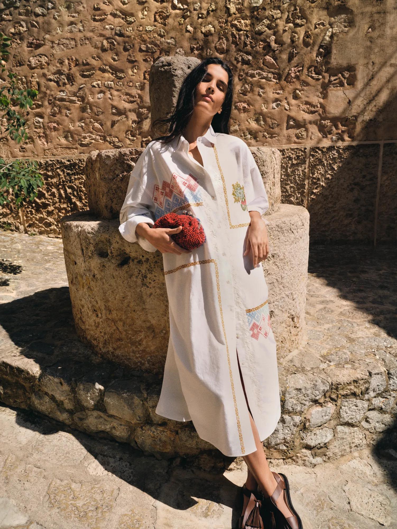 Woman in an embroidered white shirt dress with a red clutch by a historic stone fountain in sunlight