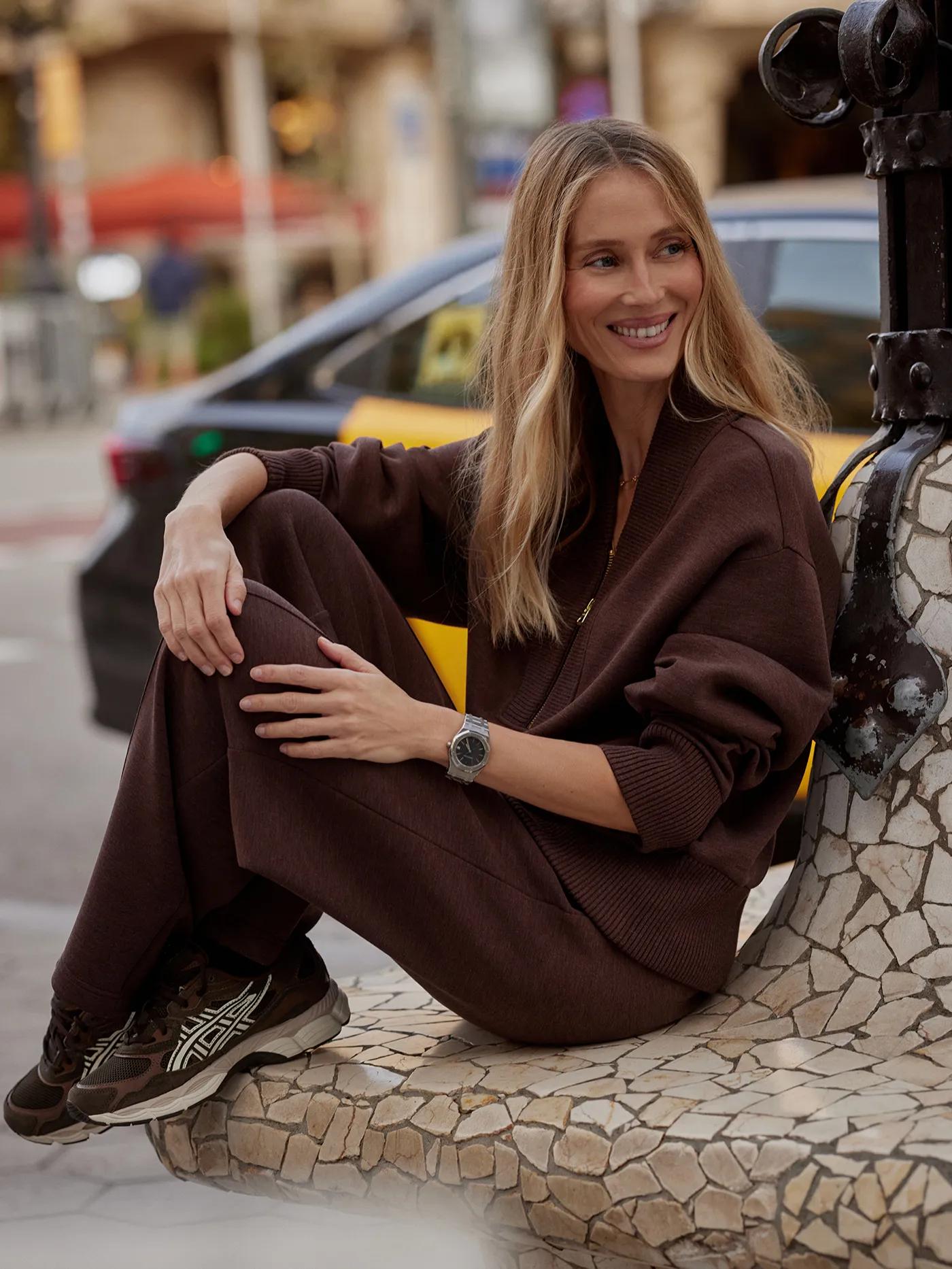 Smiling woman in a brown casual outfit and sneakers sitting on a mosaic bench in an urban setting