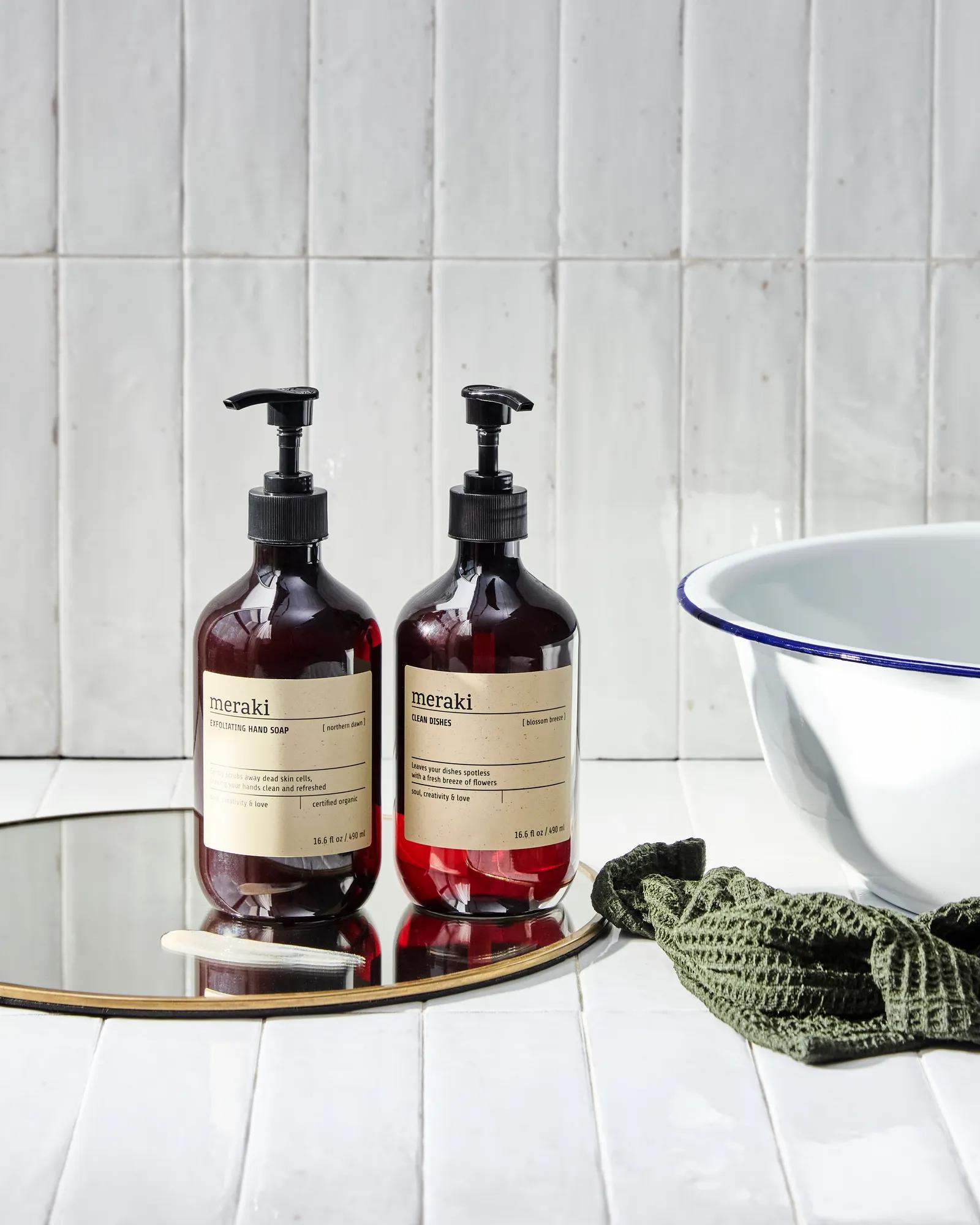 Two soap dispensers on a mirrored tray beside a washbasin and cloth against a white tiled wall