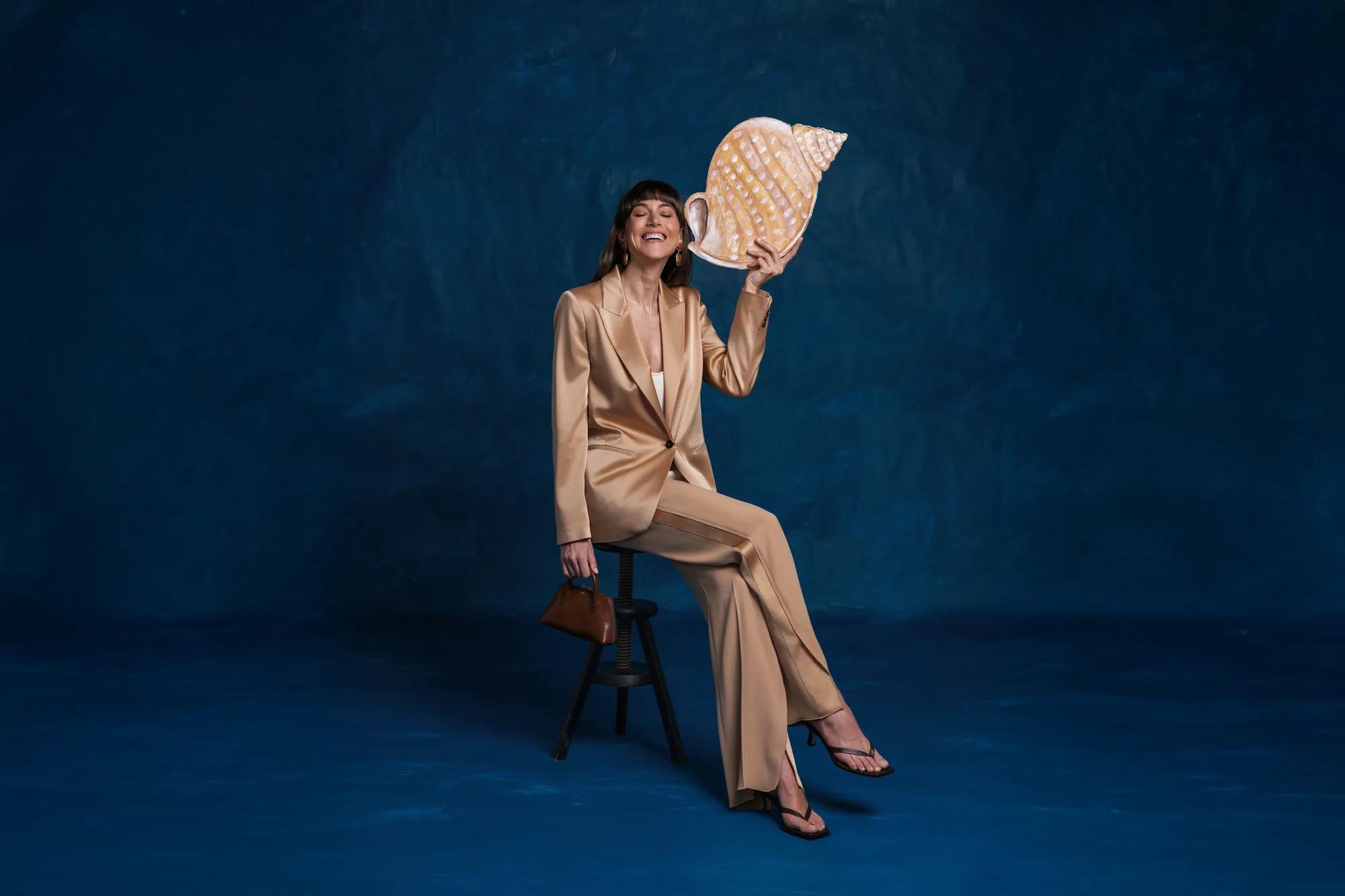 Elegantly dressed woman in a pantsuit on a stool holding a large seashell against a dark blue studio backdrop