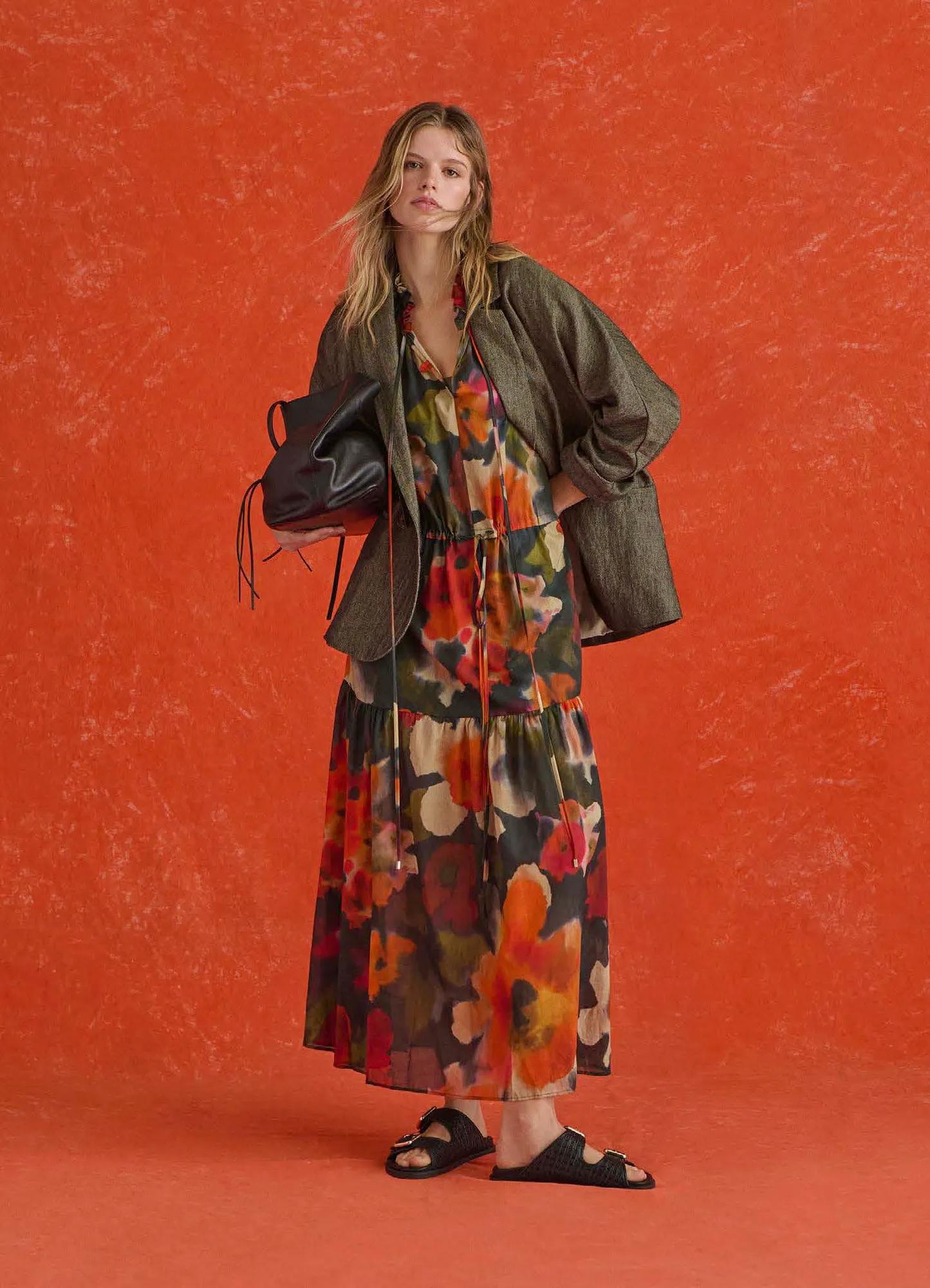 Woman in a floral midi dress with an oversized blazer and black bag against an orange backdrop