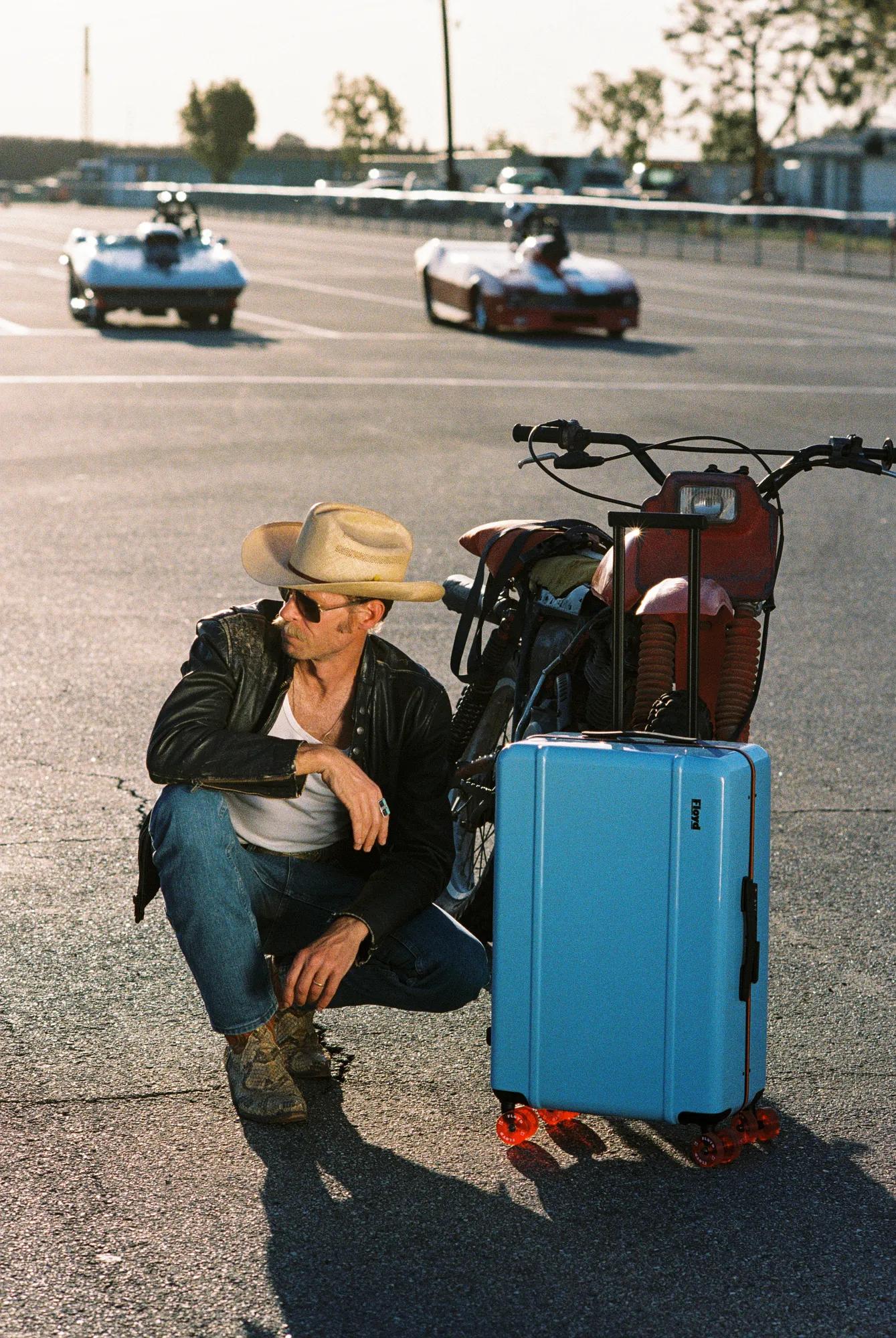 Man in a cowboy hat beside a motorcycle and blue suitcase on a racetrack at sunset