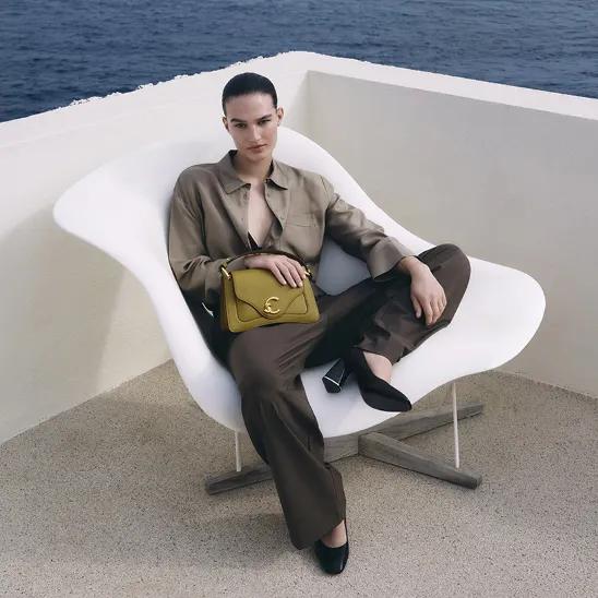 Woman in a satin two-piece outfit with a handbag seated on a modern lounge chair overlooking the sea
