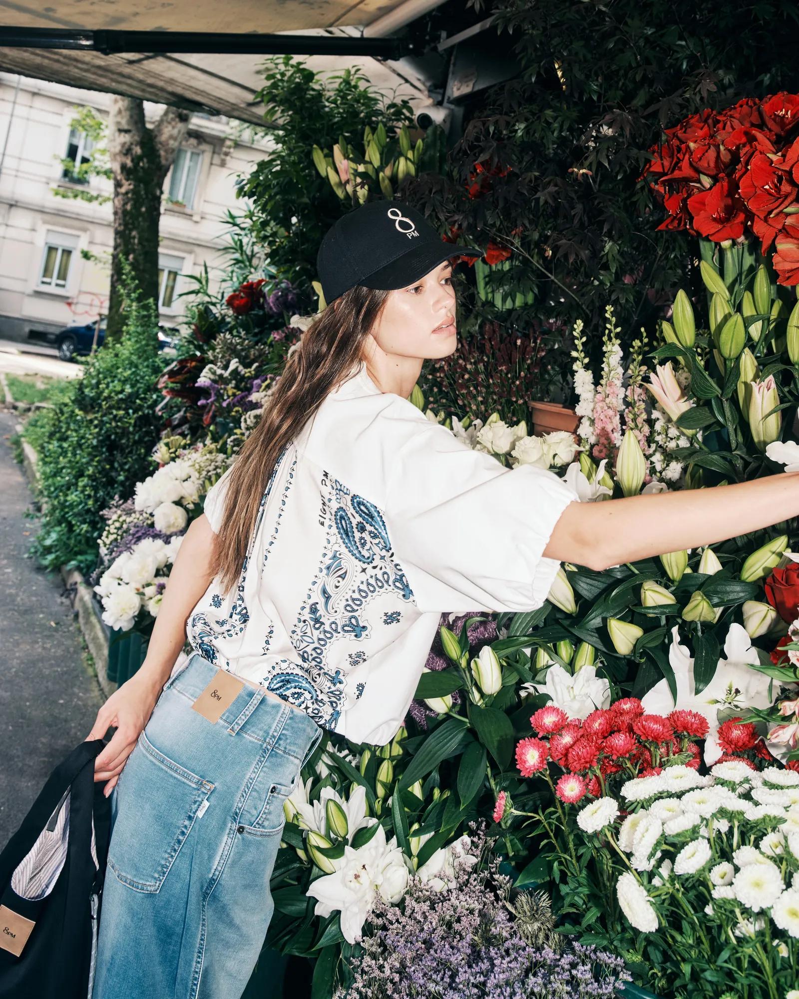 Woman in a cap, T-shirt and jeans browsing flowers at a lush outdoor flower stand