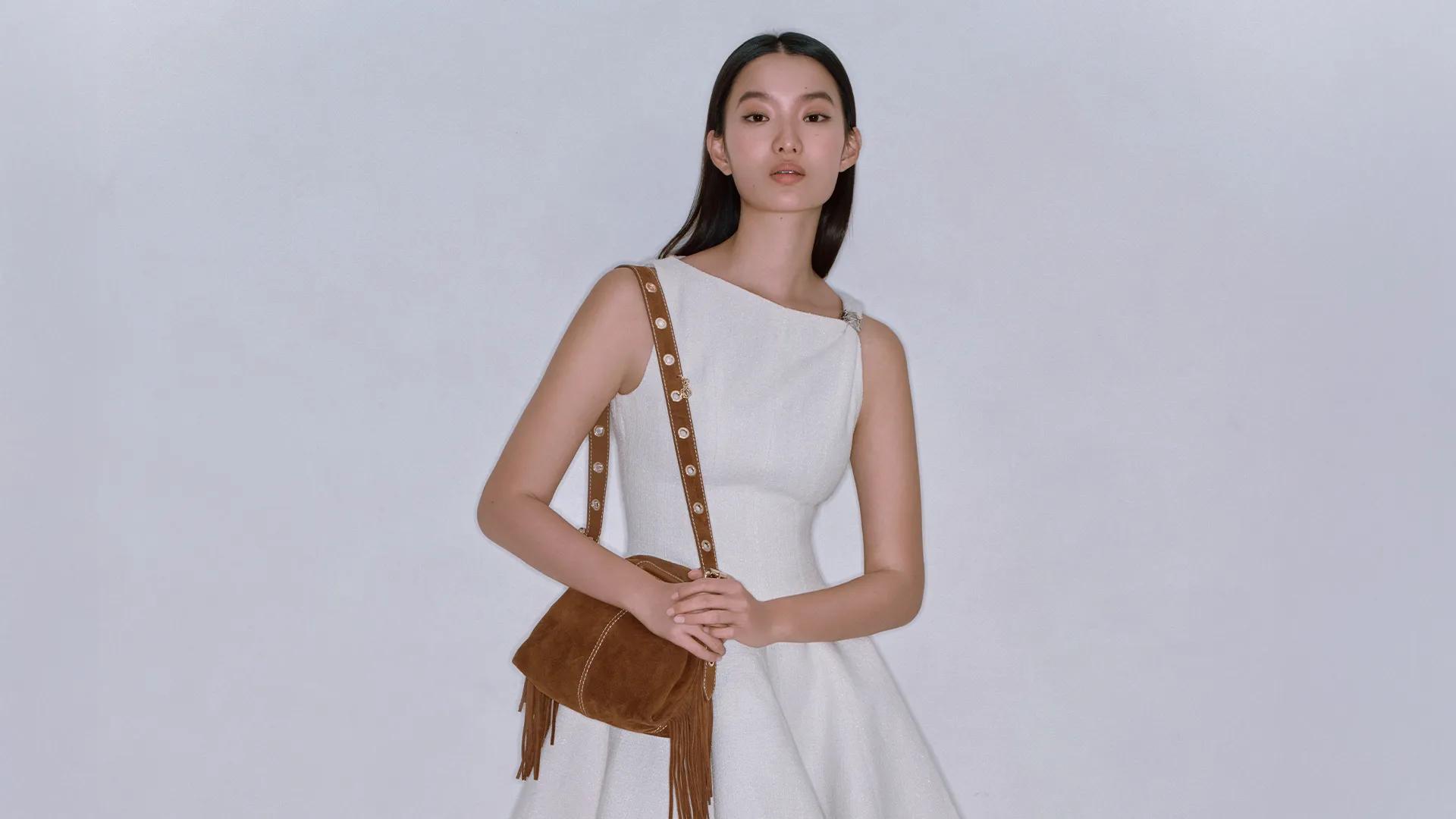 Woman in a white dress with a brown shoulder bag against a light studio background