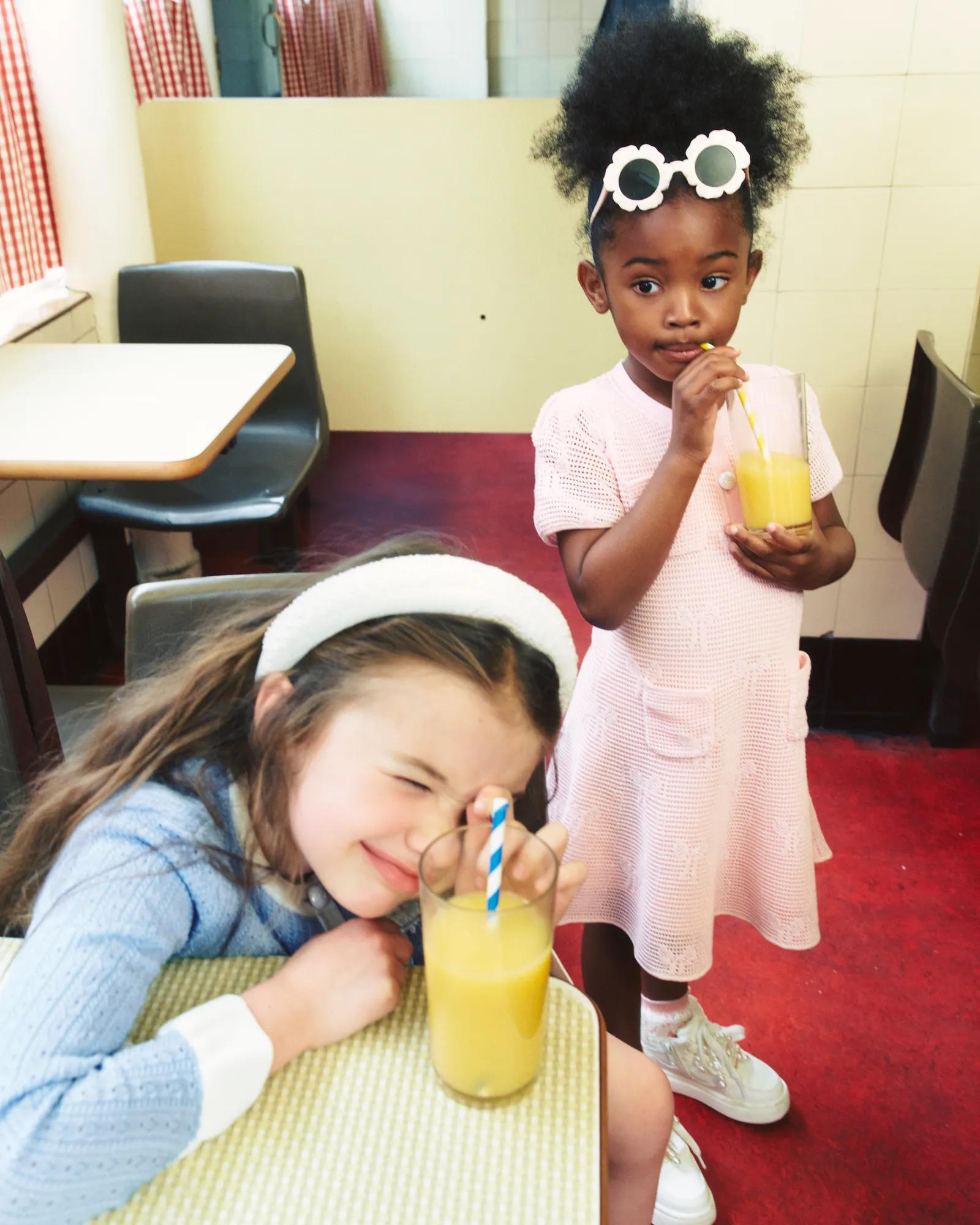 Two girls with orange juice in a café, one seated at a table and the other sipping from a glass