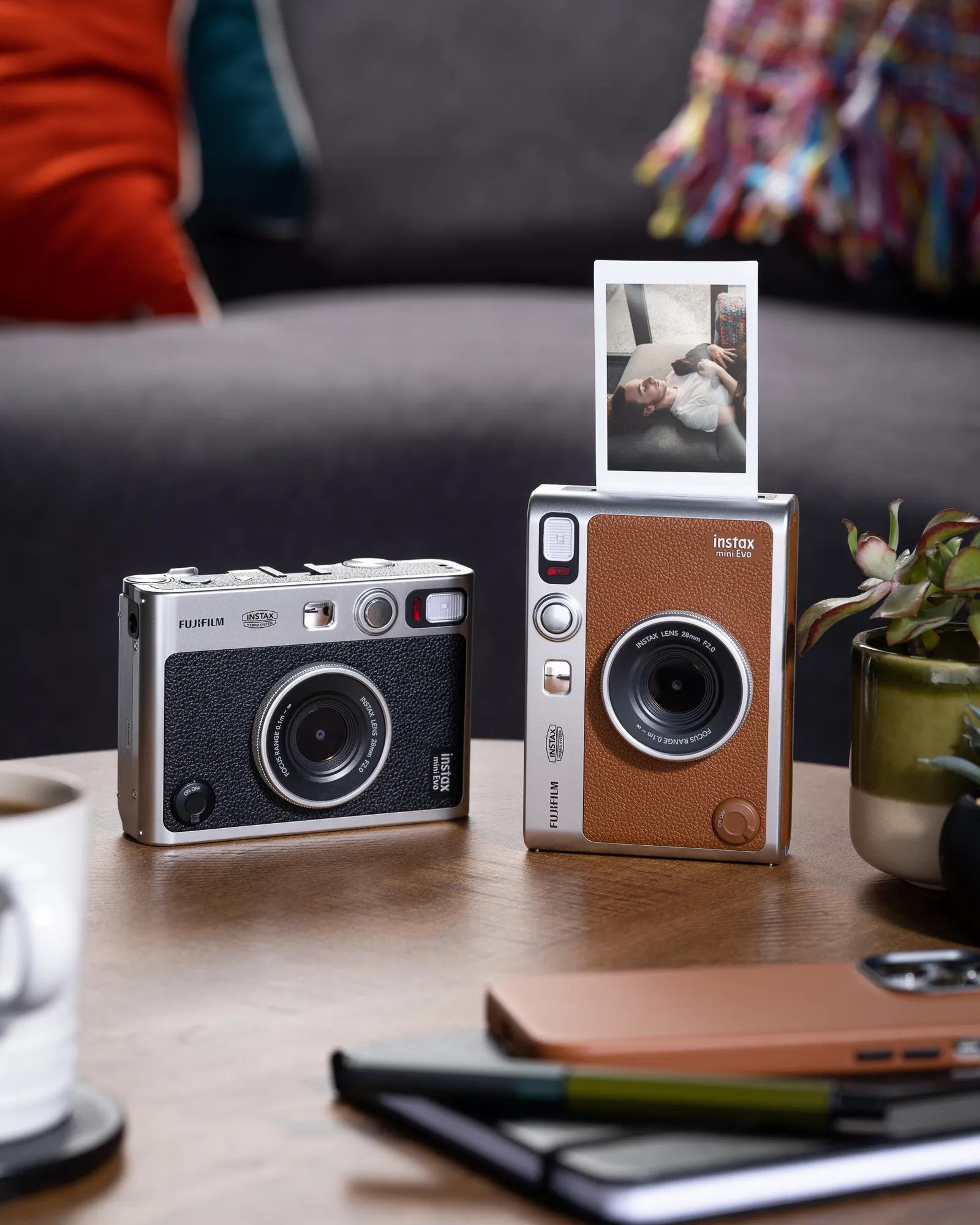 Two instant cameras on a table, one printing a photo, beside a notebook and plant