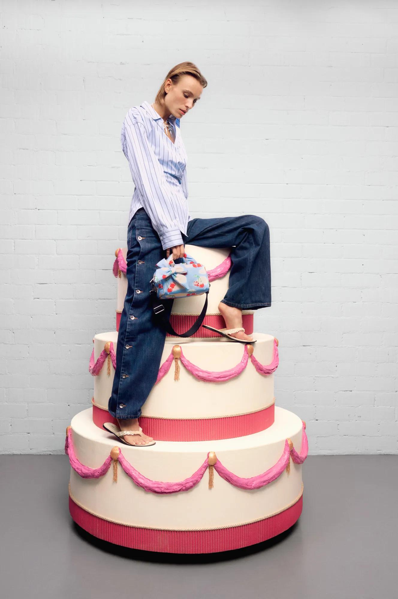Woman in a striped blouse and wide-leg jeans posing on a decorative cake while holding a small handbag
